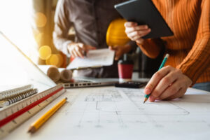 Nahaufnahme einer Hand, die auf einer Bauzeichnung arbeitet, mit einem Tablet und Bauunterlagen im Hintergrund. Perfekte Abstimmung zwischen Design und Bauausführung.