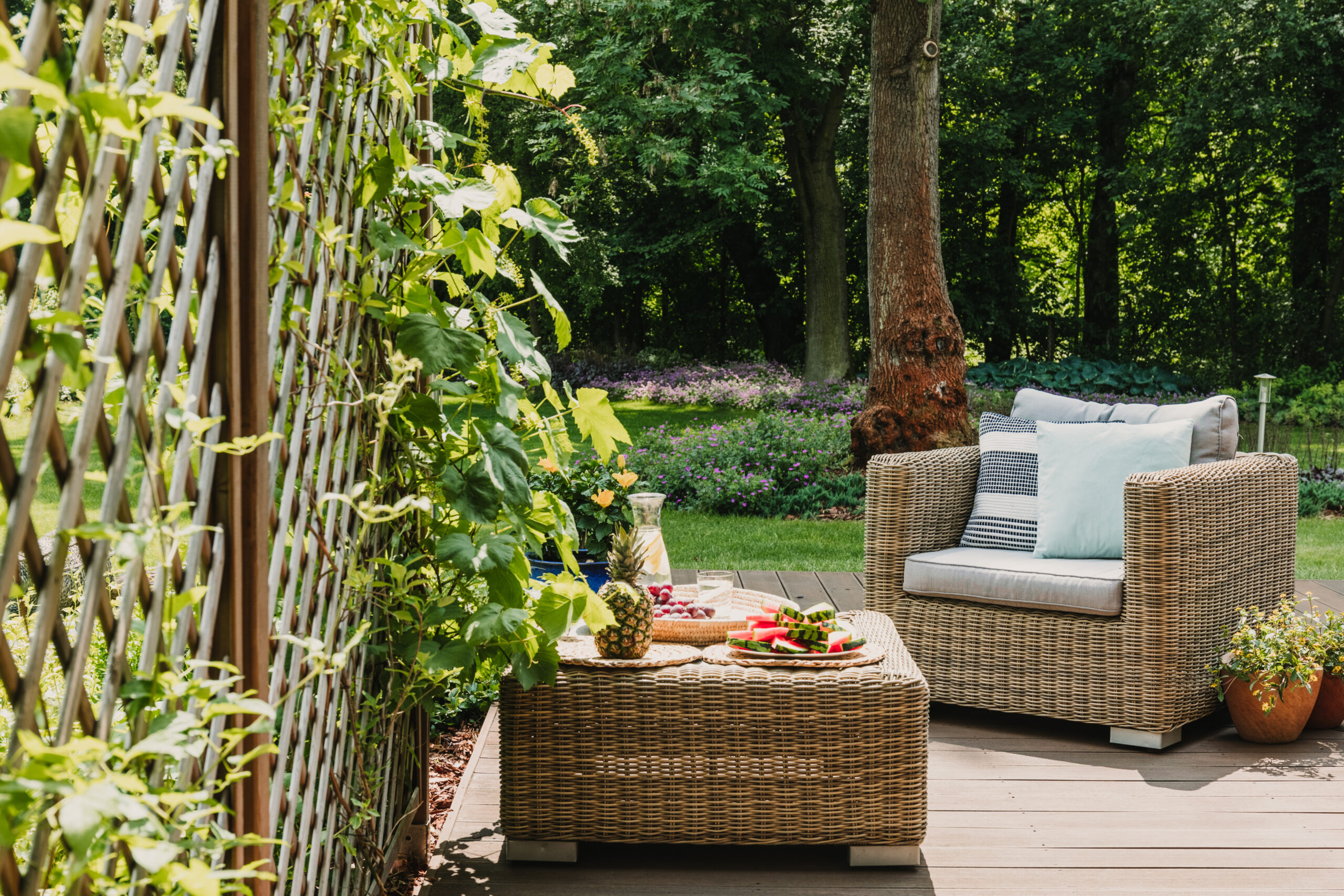Gartenterrasse mit bequemen Rattanmöbeln, Kissen und einem Tisch mit frischem Obst, umgeben von blühenden Pflanzen und grünem Rasen.